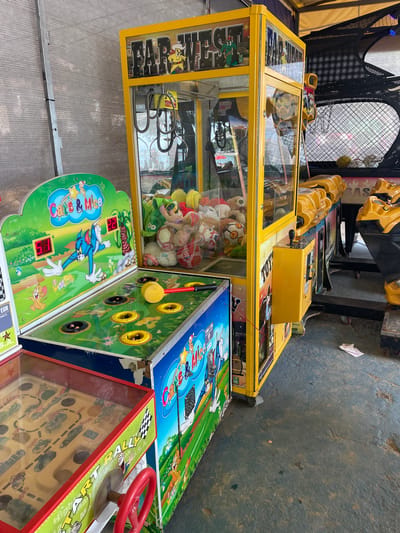 This media file shows a row of arcade games, likely in an amusement park or fairground, given the presence of crane machines and games with colorful graphics and cartoon characters. The scene is outdoors, with a textured concrete ground visible. The lighting suggests it could be daytime, possibly overcast, as there are no strong shadows.
The primary subjects are the arcade machines themselves. One prominent machine is a yellow crane game filled with plush toys. Next to it is a "Whack-a-Mole" style game featuring cartoon characters, with yellow targets and a red mallet. Further to the right is a racing game with a red steering wheel. Another machine, partially visible, has a maze-like game with small cars.
The "Whack-a-Mole" game has "Crab & Mice" written on it, along with what appear to be digital score displays showing "22" and "15". The racing game has text that includes "START", "RACE", and "GO". The overall atmosphere appears to be one of fun and entertainment, typical of an amusement setting. The location context provided is Crikvenica, Croatia, which helps to understand the setting as a public recreational area. There are no people visible in the frame. The machines appear to be operational or ready for use.