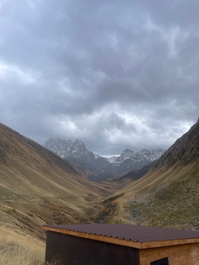 The image displays a deep mountain valley in Juta, Georgia, under a heavily overcast sky. Dense, dark gray clouds cover the majority of the sky. Two prominent mountain slopes, covered in dry, golden-brown grasses and patches of scree, frame a V-shaped valley receding into the distance. In the mid-ground and background, multiple jagged peaks are visible, several exhibiting distinct snow caps and ice patches, with one central peak partly obscured by cloud cover. The valley floor in the mid-distance contains scattered darker vegetation. In the lower foreground, the upper portion of a rectangular building is present, featuring dark brown walls, a reddish-brown corrugated metal roof with a light-colored fascia, and a small black rectangular opening on its visible right side. No people, specific actions, or interactions are depicted.