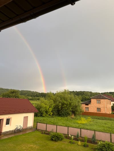 The image depicts a suburban or rural landscape with a double rainbow arching across a cloudy sky. In the foreground, a light-colored building with a burgundy roof is visible on the left, with a bicycle parked beside it. To the right, a two-story brick house with a dark roof stands behind a brown fence. The landscape is characterized by lush green grass, trees, and rolling hills in the background. The overall scene suggests a post-rain environment, indicated by the presence of the rainbow and the wet appearance of the ground and pavement. The location is identified as Hanniv, Ukraine. There are no people or animals visible in the image.