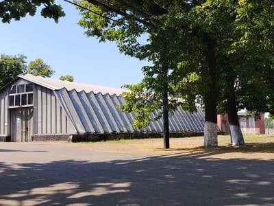 The image shows a long, grey, corrugated metal building with a distinct zigzagging roofline. The building has a large door on the left side with a window pane above it, and what appears to be a blue informational sign. The building sits on a foundation of dark brick. In front of the building is an asphalt paved area, partially covered in scattered yellow leaves, and in the foreground, large shadows cast by trees.
To the right of the building, two trees with whitewashed bases stand prominently. Further in the background, a red brick building with white-trimmed windows is visible. The sky is clear and blue. The overall scene suggests an outdoor industrial or institutional area, possibly a former industrial site or a school campus, given the style of the buildings and the presence of mature trees. The location is identified as Biaroza, Belarus. There are no people visible in the image.