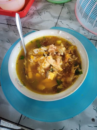 A bowl of soup, likely chicken noodle, sits on a blue plate. The soup contains noodles, chicken pieces, yellow cubes that might be potatoes or tofu, and green herbs. A silver spoon rests inside the bowl. The table underneath is gray and white with a patterned surface. Parts of a red tray and a blue and white basket are visible on the table.