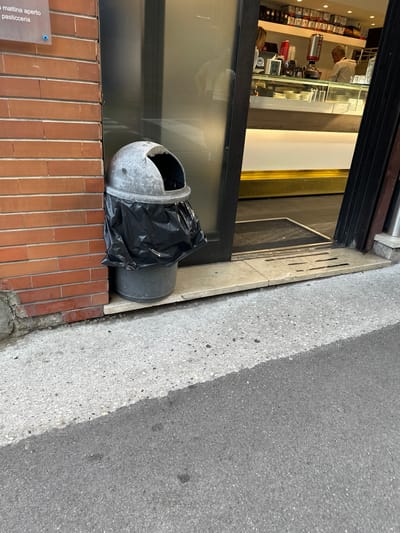 An outdoor scene shows a partially open doorway to a shop, likely a pasticceria, with a trash can in front of it. The shop interior, visible through the doorway, features a counter with a coffee machine, displays of coffee products, and two individuals, one appearing to be a customer and the other an employee. The trash can is a metal dome-shaped bin with a black plastic bag lining it, placed on a light-colored stone threshold. The building's exterior wall is made of red brick. The ground in front of the shop is paved with concrete. The image appears to have been taken during daylight hours. The shop sign above the doorway reads "La mattina aperto pasticceria," indicating it is open in the morning and is a pastry shop. While the specific location in Trieste, Italy, is not identifiable from the visual information alone, the sign's text strongly suggests a pasticceria in that city.