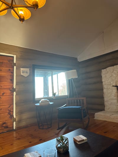 A cozy room features rustic log walls, a stone fireplace, and a wooden floor. Natural light streams through a window where a "Have a nice day" flag hangs. A floor lamp illuminates a wooden chair next to a small side table. A dark coffee table holds decorative items. A multi-bulb chandelier adds to the warm ambiance.