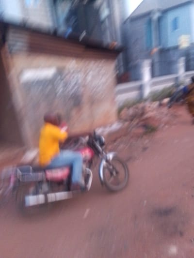 The image depicts a person in a yellow shirt and blue jeans riding a red motorcycle on a dirt road in Orlu, Nigeria. Buildings are visible in the background. The image is blurry.