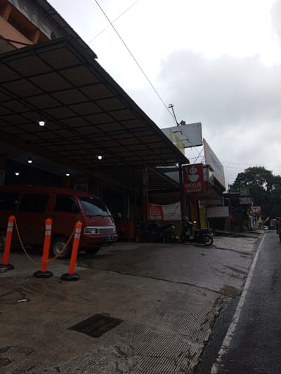 This image depicts an outdoor street scene in the city of YonPhoogla, Bhutan, under an overcast sky. The ground, consisting of textured concrete and wet asphalt, suggests recent moisture. On the left, a commercial building features an extensive, dark-paneled metal awning supported by a structural frame, beneath which several white circular light fixtures are active. A red minivan, displaying a license plate partially visible as 'L 1213...', is parked on the concrete area, preceded by three orange traffic bollards linked by a chain. A metal grate is set into the concrete surface in the foreground. Further under the awning, multiple motorcycles are parked. On the right, an asphalt road runs parallel to the concrete. Adjacent to the road, a row of commercial establishments includes a building with a prominent red rectangular sign featuring a white snow-man-like character and the word "MIXUE" in white text. A dark-colored motorcycle is parked near the "MIXUE" sign. Overhead utility lines are visible against the cloudy sky. No discernible individuals are actively engaged within the scene.