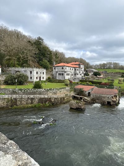 The image depicts a complex of historic stone buildings situated on the elevated bank of a flowing river in A Ponte Maceira, Spain, under an overcast sky. In the immediate foreground, the grey stone parapet of a bridge is visible on the left. The river occupies the lower third of the frame, exhibiting dark, turbulent water with white foam around various exposed rocks and smaller green vegetation clusters. A large, moss-covered boulder is prominent in the river towards the right.
On the riverbank, two smaller stone buildings with orange-red tiled roofs are visible, appearing to be old mills, with water visibly flowing from beneath the structure on the far right. Above the river, a substantial stone retaining wall supports a well-maintained green lawn.
Further up the bank, two prominent stone structures are present. To the left, a fortified-style building with crenellated walls and a dark, possibly wooden, door is set within a garden. To its right stands a larger, multi-story manor-like building, constructed of grey stone with a distinctive orange-red tile roof. This building features numerous windows, ornate stone detailing, multiple chimneys, and a grand stone staircase leading down to a formal garden with geometrically trimmed hedges.
The backdrop consists of a dense, mostly deciduous forest covering a hillside on the left. To the right, the landscape transitions to rolling green fields with scattered trees and additional smaller buildings, including a barn-like structure, under the same cloudy sky. No individuals, vehicles, or animals are visible in the scene. The overall impression is one of historical architecture integrated into a natural, rural environment.