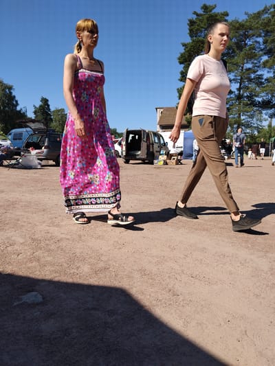 The image depicts an outdoor scene, possibly a market or fairground, with a sandy ground surface. Two women are the primary subjects. The woman on the left wears a long, bright pink dress with a floral pattern of blue, yellow, and pink circles and flowers. She has blonde hair pulled back in a ponytail and wears dark sandals. The woman on the right is dressed in a pale pink t-shirt and brown trousers. She has dark hair pulled back in a ponytail and wears black sneakers. Her gaze is directed forward, while the other woman looks slightly to her left. In the background, several vehicles, including vans, are parked. Some people, appearing to be vendors or shoppers, are visible near the vehicles and a blue tent. The overall environment suggests a sunny day, with trees and a building visible in the distance. The presence of parked vehicles and tents hints at a temporary event setup.