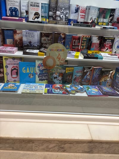 The image shows a shelf in a bookstore, likely in Calp, Spain, filled with a variety of books and children's books. The books are arranged in rows, with the spines facing outwards. Several books have prominent titles and author names visible, indicating a mix of genres, including fiction, non-fiction, and children's literature. The overall impression is one of a well-stocked book retail environment. The lighting suggests an indoor setting, possibly with natural light filtering in.