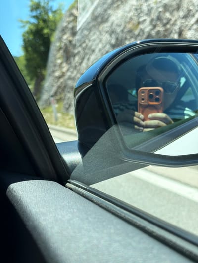 The image is a reflection in a car's side mirror, showing a person holding a smartphone. The smartphone has a prominent orange case with multiple camera lenses. The person is wearing a striped shirt and sunglasses, partially obscured by the reflection. Outside the car, a rocky hillside and green foliage are visible under a bright blue sky. The scene appears to be captured from within a moving vehicle, suggested by the angle and the slight blur of the background. The location is identified as Vrela, Montenegro.