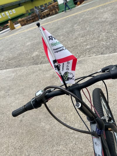 The image shows a close-up view of a bicycle's handlebars with a small flag attached. The flag is white with red trim and features text in what appears to be Korean and possibly Russian. The Korean text includes characters that might relate to safety or cycling. One part of the text on the flag reads "Helm" which is likely referring to a helmet. A cartoon image of a person wearing a helmet is also visible on the flag.
The bicycle has a bell, a gear shifter, and a cable lock secured to the frame. The background shows a paved area with yellow lines, typical of a roadway or industrial site, and a large yellow building in the distance. Pallets are stacked near the building, and a person in casual clothing is walking away from the camera in the distance. The overall scene suggests an outdoor environment, possibly near a port or industrial complex.
While the image itself does not explicitly state the location, the presence of Korean text on the flag and the industrial setting are consistent with the city of Geoje-si, South Korea, known for its shipbuilding and port activities.