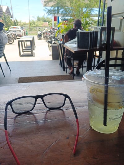 This image depicts a close-up view of a wooden table with eyeglasses and a plastic cup of beverage. The eyeglasses are black with red accents on the earpieces, resting in the foreground. To the right, a clear plastic cup filled with a light-yellow, translucent liquid and ice is visible, with slices of lemon and a black straw. A logo with the Instagram handle "@kupikeben" is printed on the cup. The background shows a blurred outdoor scene, suggesting a cafe or restaurant setting. In the distance, several motorcycles are parked, and some people are seated at tables. The environment appears to be bright and sunny, typical of an outdoor area in a tropical location. The city of Geulumpangsulu Timu, Indonesia, is noted as the location, implying a street or commercial area with vehicles and seating arrangements common in such regions.
