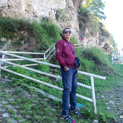 The image depicts a man standing on a sloped, grassy area with a white railing to his left. He is wearing a maroon jacket with patches on the sleeves, dark blue jeans, sunglasses, and sandals with socks. He has a dark-colored bag slung across his body. Behind him is a large, rocky cliff face covered in lush green vegetation. A small Indonesian flag on a pole is visible on the right side of the cliff. The ground in front of the railing is paved with stones and covered in grass and some debris. The overall setting appears to be an outdoor natural or semi-natural environment, possibly a park or scenic overlook.