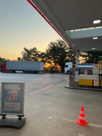 At a Shell gas station in Gorni Dabnik, Bulgaria, a red truck with a long grey trailer is parked on the asphalt. To the right, a white truck with a tanker trailer is also present. A gas pump station, marked with Shell branding, is visible on the right side of the image. In the foreground, a sign displays a special offer in Bulgarian for 2 liters of windshield washer fluid. An orange traffic cone with white stripes sits on the ground to the right of the sign. The sun is setting behind a line of trees in the background, casting a warm glow over the scene. The sky is a soft gradient of blue and orange. The overall atmosphere is that of a service area during twilight.