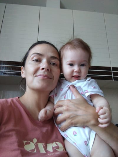 A selfie features a woman holding a baby. The woman is looking at the camera and smiling slightly, with her dark hair pulled back. She is wearing a pink t-shirt with a logo. The baby, with light brown hair, is wearing a white onesie with a strawberry pattern and is looking towards the camera with a curious expression. The background appears to be a room with white cabinets. The location is identified as Vinogradny, Russia.
