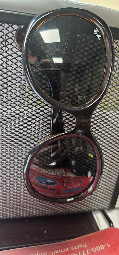This media file shows a close-up, overhead view of a pair of black Ray-Ban sunglasses resting on a black mesh surface. The sunglasses have large, oval lenses and a thick black frame with tortoise shell accents on the rims of the left lens. The Ray-Ban logo is visible in white script on the outer edge of the left lens. The lenses themselves are dark and reflective, showing a distorted reflection of what appears to be a red object with a circular logo, possibly a car emblem, and some text.
The mesh surface beneath the sunglasses is a common material used in desk organizers or ventilation grates. In the background, to the right of the sunglasses, a portion of a red object with white text is visible. The text reads "1-800-77 parts email: impo". The setting appears to be indoors, possibly on a desk or table, given the presence of what looks like a document partially visible under the mesh on the right. There are no people visible in the image. The lighting suggests it is daytime, but the indoor setting makes it difficult to determine specific time of day or weather conditions. The overall mood is neutral, focusing on the product.