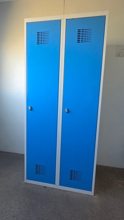 The image shows a two-door blue metal locker unit with silver trim. Each door has a silver handle, a ventilation grate near the top with a horizontal line pattern, and a ventilation grate near the bottom with a more complex pattern of horizontal and vertical lines. The locker unit stands on a dark gray carpeted floor. The background consists of a white wall on the left, illuminated by a light source casting a shadow, and a light gray wall on the right, with a white electrical outlet visible. The scene appears to be indoors, in a room that could be a changing room, school, or office environment. There are no people or specific actions depicted. The location is not specified within the image itself.