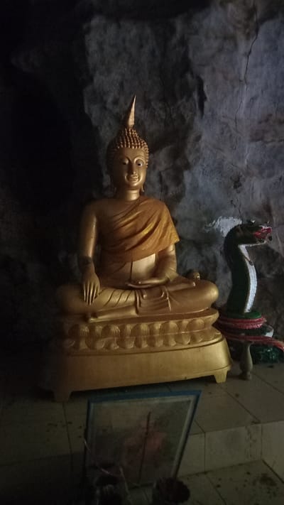 A golden Buddha statue sits in a cave temple in Ban Pak Ra, Thailand. The statue is resting on its side. It is adorned with a golden robe and has a pointed topknot. Above the statue is a colorful serpent figure, possibly a naga. To the side, there is a framed image leaning against a wall. The walls of the cave are rough and dark.