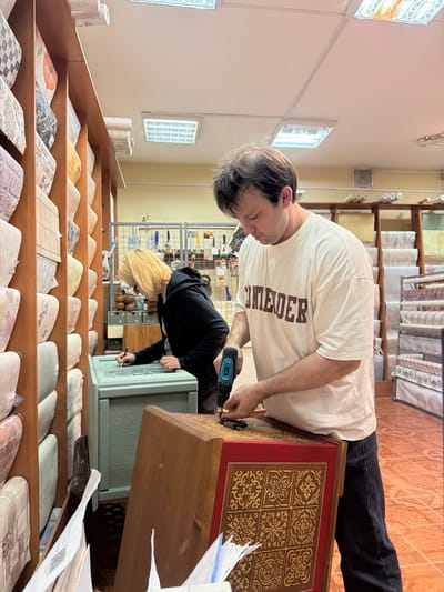 In Maloyaroslavets, Russia, two individuals are engaged in work within a shop, possibly a fabric or wallpaper store, indicated by the numerous rolls of material displayed on wooden shelves lining the left side of the frame. In the foreground, a man wearing a cream-colored t-shirt with maroon lettering that reads "CONTENDER" and dark jeans is using a cordless drill to attach something to a red-and-gold decorative wooden box. His focus is on the task at hand. To his left, a woman with short blonde hair and wearing a black hoodie is bent over a light green chest or cabinet, appearing to be drawing or marking on its surface with a pen. The chest has an embossed or carved decorative pattern. The floor is tiled with orange and brown square tiles. In the background, more shelves with rolls of material are visible, along with some tools and various items stacked on shelves. The lighting suggests an indoor commercial space.