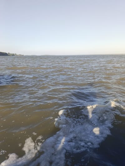 The image depicts a body of water, likely a lake or a large river, under a clear sky. In the foreground, there is foamy water, suggesting movement from a boat or disturbance. The water is a murky brown color, with ripples and small bubbles visible. In the distance, a tree line and a faint horizon are discernable. The location is identified as Krasnyi Yar, Russia. The scene is a natural outdoor environment with no visible people or specific man-made objects other than possibly a submerged structure causing the foam in the foreground.