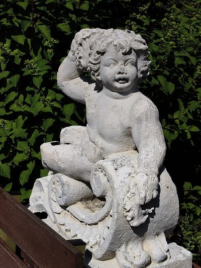 This media file is a close-up, outdoor photograph of a weathered, white stone statue of a cherubic child. The statue is seated on a decorative base that appears to be carved to resemble a scroll or a wave. The child has curly hair, large eyes, and an open mouth, giving it a lively and somewhat surprised expression. Its right arm is raised, with the hand resting on its head, while its left arm is bent at the elbow.
The statue is set against a backdrop of lush, dark green foliage, suggesting a garden or natural setting. The sunlight highlights the texture of the stone, revealing cracks and peeling paint, which indicate age and exposure to the elements. The lighting suggests it is daytime, with strong shadows cast by the statue and the surrounding leaves. In the foreground, a section of a wooden fence or railing, made of dark brown planks, is visible, adding a rustic element to the scene.
There is no visible text in the image. The overall mood is one of stillness and artistic observation, with the ancient-looking statue juxtaposed against vibrant, living greenery. The location context of Goczałkowice-Zdrój, Poland, implies this is likely a decorative piece in a garden or park within that region. The statue itself is the primary subject, with the natural environment serving as its setting.