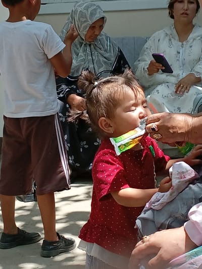 This is a medium shot photograph taken outdoors in Osh, Kyrgyzstan. The scene appears to be a family gathering. In the foreground, a young child in a red polka-dotted dress is being offered something from a wrapper by an adult hand adorned with rings. The child's eyes are closed, and the wrapper is partially in their mouth. To the left, a young boy in a white t-shirt and brown shorts stands facing away from the camera. In the background, two women are seated. One woman, wearing a patterned headscarf and dark clothing, looks down. The other woman, dressed in a white floral patterned outfit, is holding a purple mobile phone and looking forward. The ground is paved and sunlit, with shadows suggesting an outdoor setting.