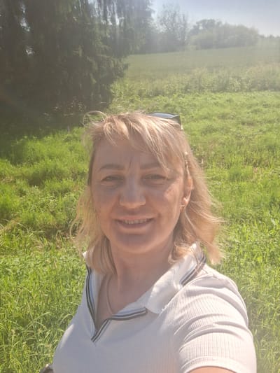 A woman with blonde hair and a white collared shirt is taking a selfie outdoors. She is smiling and has sunglasses on top of her head. The background is a grassy field with trees in the distance. The sun is shining brightly, creating a lens flare effect in the image. The overall scene suggests a casual, daytime setting in a natural environment.