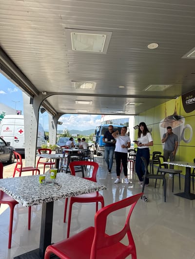 The image shows a group of people standing and sitting at tables outside a cafe or rest stop. The scene appears to be a sunny day, with a bright sky and some clouds visible in the background, along with mountains. The setting is an outdoor area with a covered roof, likely a roadside cafe or a service station.

There are several people visible. Some are standing, appearing to converse or wait. Others are seated at tables. One group of three men is gathered around a table under the awning, with one man leaning over the table and the other two standing behind him. Another woman is standing, looking towards the camera, while a man in a gray shirt is visible in the background, also appearing to be engaged in conversation.

Objects in the scene include several red chairs and black chairs, as well as tables with patterned tablecloths. There are also cups and a glass ashtray on one of the tables. In the background, vehicles are visible, including a white delivery truck with text that includes "DOSTAL" and "LIJEŠNJE", and a blue truck. There's also a glimpse of a gray car.

The overall mood seems relaxed and casual, possibly a stop during a road trip or a break at a service area. The time of day appears to be daytime, given the bright lighting. The location context provided is Liješnje, Montenegro, and the text on the truck supports this. A large graphic on the wall behind some of the people features a coffee cup and the text "FRESH CORNER".