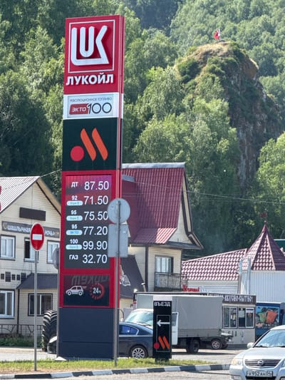 The image shows a gas station with a prominent Lukoil sign in Mayma, Russia. The sign displays prices for various fuel types including diesel (ДТ), gasoline (92, 95, 100), and gas (ГАЗ). The prices are 87.50, 71.50, 75.50, 77.20, 99.90, and 32.70 respectively. There is also an advertisement for "Revolutionary Fuel" called "EKTO 100".
The gas station appears to be part of a larger commercial area, with buildings visible in the background. One building has a sign in Russian that appears to say "Shashlychnaya" (a type of fast food eatery) and another sign that reads "Night". Another building has the text "ВКУСНО-СЫТНО!" which translates to "Tasty-Satisfying!".
A truck with a grey tarp and a sign indicating "ВЪЕЗД" (Entrance) with an arrow pointing left is parked near the gas station. A black car is also visible. In the distance, a rocky hill covered in trees is visible, with a flag at its summit. The overall environment is sunny, with green trees and foliage. The ground in the foreground is paved with a white and black striped curb.