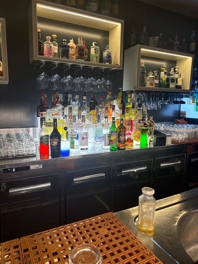 The image depicts a well-stocked bar counter in what appears to be a commercial establishment. The counter is made of stainless steel and is lined with a variety of liquor bottles, including brands like Baileys, Absolut, and various vodkas and gins. There are also numerous stacks of clean drinking glasses and wine glasses hanging upside down on racks above the counter. Behind the counter, shelves are illuminated and display more bottles of alcohol. To the left of the counter, a digital display shows "88.6" and below it, a large numeral "5" is visible on a cabinet. In the foreground, a brown grid mat is placed on the counter, and a small, empty glass bottle with a white cap sits near a sink. The overall environment suggests a functioning bar ready for service. Without additional context or visual cues, it is not possible to definitively ascertain the specific location within Giugliano in Campania, Italy.