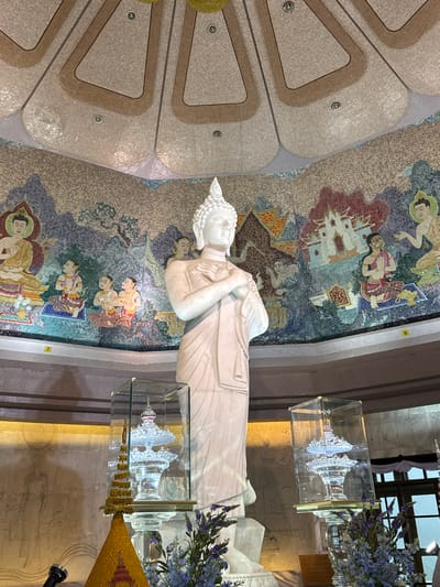 The image shows the interior of a temple in Chang Khoeng, Thailand, focusing on a large white marble statue of a standing Buddha. The Buddha statue is positioned centrally and is depicted with clasped hands. Behind the statue, the curved walls are adorned with intricate mosaic art depicting various scenes and figures, characteristic of Buddhist art. The ceiling above is also decorated with a mosaic pattern and features recessed lighting.
In front of the Buddha statue, to the left and right, are glass display cases. The case on the left contains a golden tiered structure topped with a crystal-like ornament. The case on the right also contains a tiered crystal-like ornament. In the foreground, a microphone is visible on the left, and a golden triangular structure with red accents is present. Blue and purple flowers are arranged in vases on either side of the display cases, adding to the decorative elements of the space. The overall environment suggests a sacred and artistic place of worship.