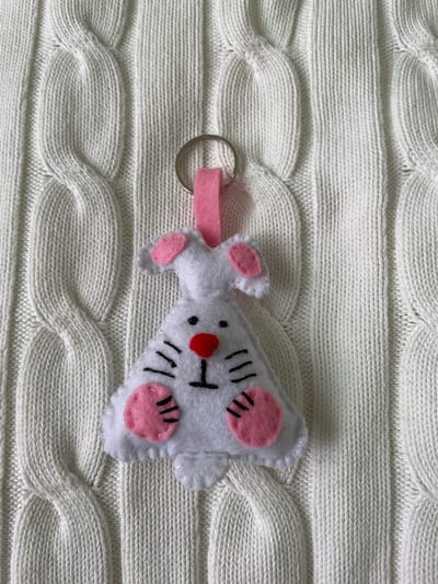 The image shows a handmade felt keychain of a white bunny with pink details on a white cable-knit sweater. The bunny has a red nose and black whiskers. The keychain has a silver ring and a pink felt loop. The background is the texture of the sweater. The location is Лесной Пейзаж Кп (шарапово, Russia is not evident from the image.