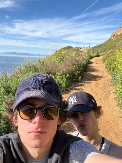The image displays a selfie taken by two individuals on a coastal dirt trail in Palos Verdes Estates, United States, under a clear, sunny sky. The foreground is dominated by a young adult male on the left, wearing a navy blue corduroy baseball cap with an "LA" logo, and dark sunglasses with round amber-tinted lenses over a black frame. He is dressed in a dark grey hooded top over a light grey t-shirt, looking directly at the camera with a neutral expression. Partially visible to his right is a young adult female, wearing a dark blue baseball cap with a white "NY" logo and a light grey ribbed tank top. She is puckering her lips towards the camera.

The background features a wide dirt path winding uphill to the right, bordered by green shrubs and patches of yellow wildflowers. Several indistinct figures, consistent with hikers, are visible further up the trail. To the left, the Pacific Ocean extends to the horizon, where a hazy landmass, identifiable as Catalina Island, is visible with a few distant cargo ships. The sky is bright blue with scattered white clouds and a faint aircraft contrail. The scene is brightly illuminated by direct sunlight.