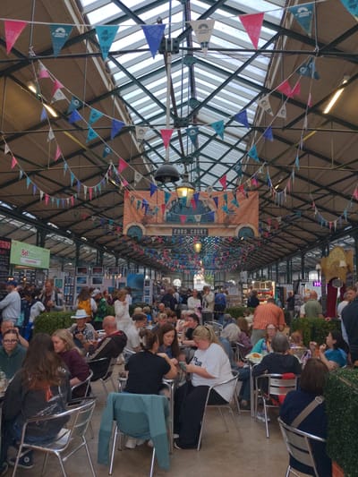 This is an interior shot of a bustling food market in Belfast, United Kingdom, likely St. George's Market, given the prominent St. George's Market branding on the bunting. The market is covered by a large, arched, glass and steel roof structure, allowing natural light to filter in. Rows of stalls and food vendors are visible, with people milling about and seated at tables. The scene is decorated with numerous strings of colorful triangular bunting, featuring the St. George's Market logo in white, as well as plain colored flags in pink, blue, and teal. In the background, a large banner reads "FOOD COURT." People are engaged in various activities: eating, talking, shopping, and observing. The overall atmosphere appears lively and crowded, with a mix of adults and some younger individuals. Several food stalls are evident, including one advertising "Bel Falafel" and another with a sign for "Crêperie." The architectural style suggests a historic market hall.