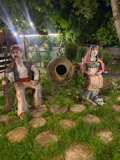 The image captures a scene in Verin Ptghni, Armenia, featuring a garden or yard setting. There are two statues of people depicted in traditional Armenian clothing along with an earthen jar sitting on a metal holder. One statue represents a woman wearing a headdress and traditional attire while seated next to a sculpture of a cat. The other statue is that of a man holding a rifle. Both statues appear to be positioned alongside a path consisting of wooden discs set into the grass. In the background, there is a cage-like structure surrounded by trees, suggesting an outdoor, perhaps park-like environment. The overall setting implies a cultural or traditional decorative display within a garden.