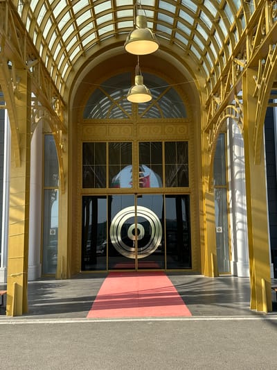 An ornate, golden architectural entrance stands prominently, featuring a grand arched canopy and a red carpet leading to the main doors. The structure, likely a commercial or public building in Kapitan Andreevo, Bulgaria, is illuminated by bright daylight, casting distinct shadows. The arched canopy overhead is composed of golden latticework and translucent panels, resembling a skylight. Two large, golden pendant lights hang from the canopy's ceiling. The facade is predominantly golden, with decorative elements, including circular motifs above the central doors and intricate scrollwork on the supporting arches and balustrades. The main entrance consists of double glass doors, dark and reflective, featuring a large, stylized golden concentric circle logo on the right door. Large arched windows above the doors and rectangular windows to the sides also have golden frames. Reflections in the glass show a landscape with hills and trees, vehicles, and a flag with horizontal red, white, and green stripes. A bright red carpet runner extends from the doors across a dark grey tiled pavement towards the foreground. A narrow asphalt road with a white line is visible at the very bottom of the image. No individuals are present in the immediate vicinity of the entrance.