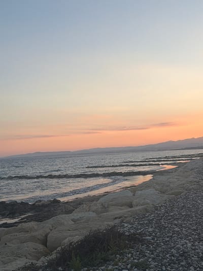 A wide shot captures a serene beach scene at dusk in Pervolia, Cyprus. The sky transitions from a pale blue at the top to soft hues of orange and pink near the horizon, with faint clouds adding subtle texture. A range of distant, hazy mountains forms a silhouette against the horizon. The sea, a muted blue-gray, features gentle waves lapping at the shore, marked by a series of rocky breakwaters. The foreground of the beach is a mix of large, pale rocks and smaller dark pebbles, with a sparse scattering of dry vegetation. The overall atmosphere is tranquil and natural.