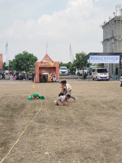 The image captures an outdoor event in Plosorejo, Indonesia, featuring a group of children engaged in a playful activity on a dry, grassy field. Two boys are at the center, one appearing to be tackling or wrestling the other who is on the ground. Both boys are wearing casual attire. In the background, a banner that reads "POSKO BANSER" with smaller text below indicates a local community post or event. Adjacent to this is an ambulance with "AD 0642 XV" on its license plate. Further back, a stage-like structure with "DJARUM 76" branding and people gathered around it suggests a larger celebration or festival. Several vehicles, including cars and motorcycles, are parked in the vicinity. The sky is partly cloudy. The overall scene depicts a lively community gathering with recreational activities for children.