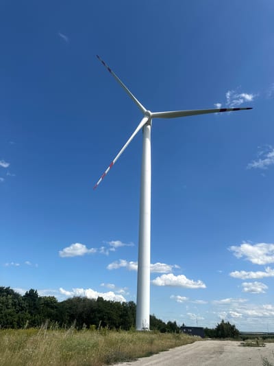 A tall, white wind turbine stands against a bright blue sky with scattered white clouds. The turbine has three blades, each marked with alternating red and white stripes near the tip. The turbine is positioned on a slight incline with dry grass and weeds in the foreground. A dirt road runs diagonally from the lower right corner towards the center of the image. In the background, a dense line of dark green trees is visible, and beyond that, a low-lying building with a dark roof can be seen. The scene appears to be in an open, rural area under a clear sky.