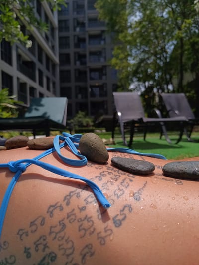 A close-up shot features a person lying outdoors, presumably sunbathing, with a blue bikini top partially visible. The person's back is adorned with tattoos written in a foreign script. Several smooth, rounded stones of varying sizes and colors (brown and gray) are arranged on the person's back. Water droplets are visible on the skin.
The background is blurred, but it appears to be a resort or apartment complex with lush green vegetation. Several lounge chairs are visible. The photo was taken in Hua Hin City Municipality, Thailand.