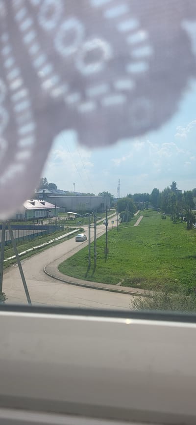 This is a photograph taken from a window, with a patterned fabric obscuring the top left portion of the image. The view is of a rural or semi-urban environment, likely in Gamovo, Russia, under a partly cloudy sky.
In the foreground, a white or light-colored windowsill is visible at the bottom of the frame. Beyond the windowsill, a paved road curves from the left side of the image towards the center. On the road, a silver car is visible, moving away from the viewer. Several utility poles with wires run along the side of the road.
To the left of the road, there is a green fence and a building with a green facade and a dark roof. Further back, there is a larger, flat-roofed building, likely an industrial or commercial structure, with several parked cars in front of it. A few smaller houses with pitched roofs are also visible behind the green building.
To the right of the road, there is a large expanse of green grass and trees. In the distance, a tall metal tower, possibly a communication tower or electricity pylon, is visible against the sky. The overall impression is one of a quiet, functional area on a summer day.