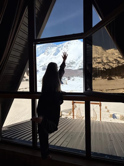 An indoor view through a large, multi-pane window reveals a snow-covered mountainous landscape under a clear blue sky in Virak, Montenegro. A person, observed from behind in silhouette, stands facing the window. The individual, with long hair, is dressed in dark clothing, consistent with loungewear or pajamas. Their right arm is raised, hand extended palm-forward towards the distant snow-covered mountains. Their bare left foot is elevated, resting on a horizontal window frame or sill.

The interior structure features dark window frames and a visible sloped ceiling on the left, indicating an A-frame architectural style. Outside the window and directly below, a dark, corrugated metal roof is partially visible. Beyond the roof, a snow-covered expanse shows visible tracks and leads to an outdoor seating arrangement featuring two suspended, partially snow-covered, woven chairs hanging from a wooden frame. The mid-ground includes a valley with scattered small buildings and evergreen trees. The background is dominated by large, extensively snow-covered mountains. The scene depicts a clear, sunny winter day.