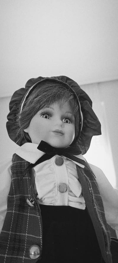 A black and white, low-angle, close-up shot captures a porcelain doll wearing a bonnet and period clothing. The doll's eyes are wide open, and it has long eyelashes. It is dressed in a white ruffled blouse with a black bow tie, a plaid vest, and a dark garment that appears to be a skirt or pants. The doll is positioned indoors, against a plain, bright background. The image is grainy. The location is not visible in the image.