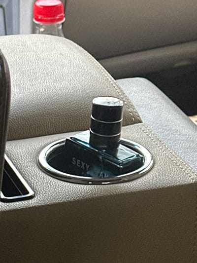 The scene depicts a detailed view inside a vehicle, located in Huangdao District, China. A rectangular glass perfume bottle with a dark, cylindrical cap rests within a chrome-rimmed cup holder. The bottle, which appears to contain a light blue liquid, prominently displays the word "SEXY" and partial text "AM" on its side. The cap is dark, possibly black, and features three distinct horizontal silver bands. The cup holder is integrated into a padded center console or armrest, upholstered in a light beige or gray material with a finely textured surface. In the blurred background, a transparent plastic bottle with a red screw-top cap is partially visible, positioned behind the console. No individuals are present in the image, and no active interactions are occurring.