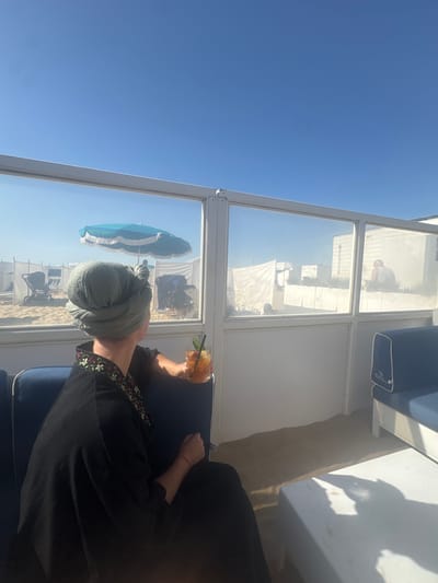 The image shows a person wearing a head wrap and dark clothing, seated on a blue upholstered chair inside a structure with window-like partitions. The reflection in the glass reveals a beach scene with beach umbrellas, chairs, and people. A cocktail is visible in the person's hand. The location is Knokke-Heist, Belgium.