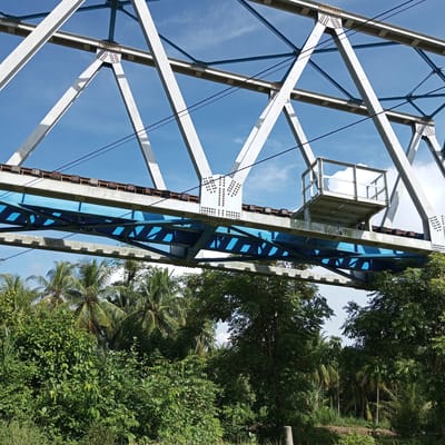 This media file captures a low-angle view of a steel truss bridge spanning across a lush, green landscape. The bridge, painted in white and blue, features a complex network of beams and supports, with railway tracks visible on its upper deck. A small metal platform with railings is attached to the side of the bridge, near the center. Below the bridge, dense vegetation, including palm trees and various other leafy plants, dominates the scene. The sky above is a clear blue with a few scattered white clouds.
The image appears to have been taken during the daytime, as indicated by the bright sunlight casting shadows on the bridge structure and illuminating the foliage. The weather seems pleasant, with clear skies and good visibility. The location context suggests this scene is in Blangkeude, Indonesia, which is consistent with the presence of tropical vegetation. No people are visible in the image, and there are no apparent activities or events taking place.
A notable detail on the side of a white beam on the bridge is a pattern resembling the letters "TM" formed by small rivets or bolts. This could be a designation or part of the bridge's identification. The overall impression is one of a functional piece of infrastructure integrated into a vibrant natural environment.