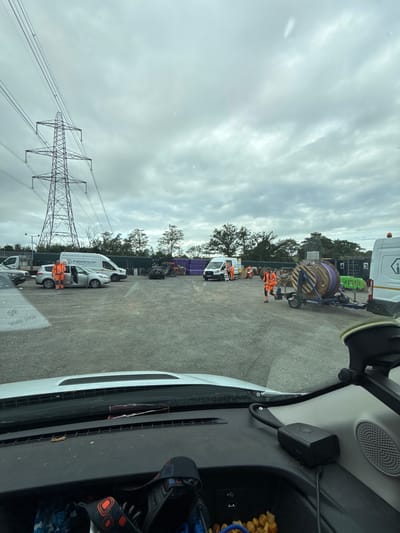 In an outdoor industrial setting, under a cloudy sky, a utility worker in an orange hi-vis jacket and trousers stands next to an open silver car door. Several vehicles, including vans and cars, are parked on a gravel lot. A large electrical pylon is visible in the background, along with trees and fencing. In the mid-ground, a large wooden spool, likely containing cable, is mounted on a trailer. Other workers in similar orange attire are present, some appearing to be engaged in work related to the spool. The scene is viewed from the interior of a vehicle, looking out through the windshield.