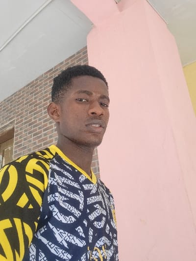 A young male individual, appearing to be in his late teens or early twenties, with dark skin and short black hair, is depicted in a close-up, chest-up portrait. He wears a short-sleeved t-shirt featuring a complex graphic pattern: the sleeves and upper shoulders are black with large yellow abstract shapes, while the main body is black with intricate white abstract symbols and lines. A yellow collar trim is visible. The subject is looking directly towards the viewer with a neutral facial expression. The scene is located in Ebenebe, Nigeria. Behind the subject to the left, a reddish-brown brick wall is visible, along with a partial view of a window frame or wooden structure. To the right, a large, light pink rectangular pillar or wall section dominates the background. A light grey or white ceiling or overhang is visible above the subject's head. The lighting is bright and even.