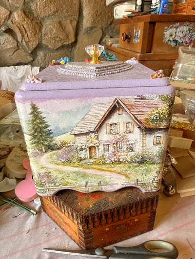 The image displays a decorative lidded box, painted with a pastoral scene of a cottage, a winding path, and trees, sitting atop another smaller, intricately carved wooden box. The lid of the larger box is adorned with a jeweled embellishment and delicate floral accents. The background consists of a stone wall and various craft supplies, suggesting a workshop or crafting area. The scene is lit with natural light, casting subtle shadows. There are no people present in the image, and no specific location in Slavyani, Bulgaria can be identified from the visual information provided.