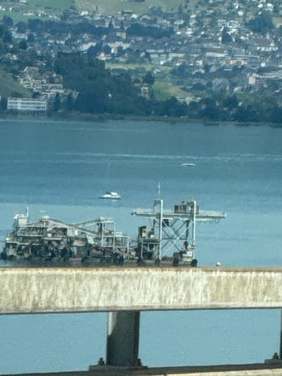 The image depicts a scene with a body of water, a floating structure, and a town on a hillside in the background. The floating structure appears to be a pontoon or barge with a complex arrangement of metal scaffolding, platforms, and possibly machinery. It is positioned in the middle ground of the image. Further in the distance, on the water, two small boats are visible. The background shows a densely populated town built on a steep, green hillside, characteristic of towns located near lakes in mountainous regions. The architecture of the town includes numerous buildings with light-colored roofs and walls, interspersed with trees. The overall impression is of a scenic landscape, likely a Swiss town on a lake. Specifically, the town is located in Beckenried, Switzerland, which is situated on the shores of Lake Lucerne and surrounded by the Swiss Alps. The foreground of the image features a part of a concrete or stone structure, possibly a pier or a bridge railing, running horizontally across the bottom half of the frame, partially obscuring the view of the water and the floating structure. The water itself is a calm, blue expanse. The lighting suggests it is daytime, with soft natural light.