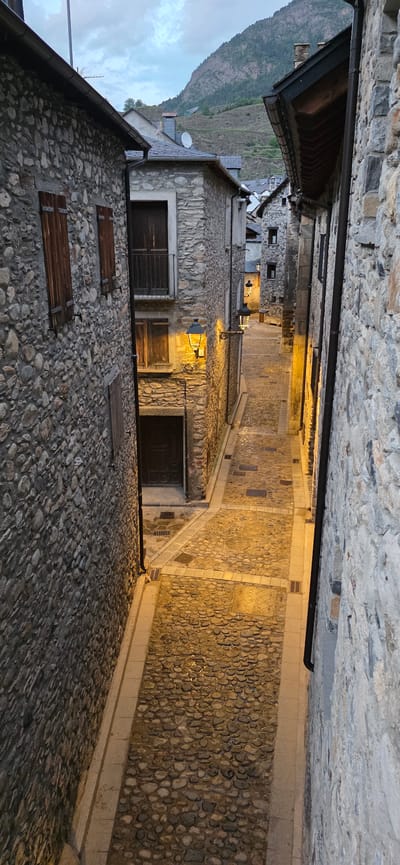 The image shows a narrow street in Benasque, Spain, flanked by stone buildings. The street is paved with cobblestones and lit by yellow streetlights. The buildings are constructed from roughly hewn stone, with dark wooden doors and shutters. A mountain is visible in the background under a dusky sky. The buildings appear old and well-maintained, with visible stone variations and textures.